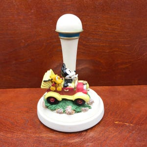 May include: A white ceramic figurine of Mickey Mouse and Pluto driving a red car. The figurine is on a white base with a green grass and brown rock design. The figurine is a decorative piece for a home or office.