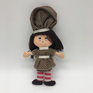 May include: A brown and white polka dot doll with brown hair, wearing a white and brown dress, white and brown striped stockings, and black shoes.