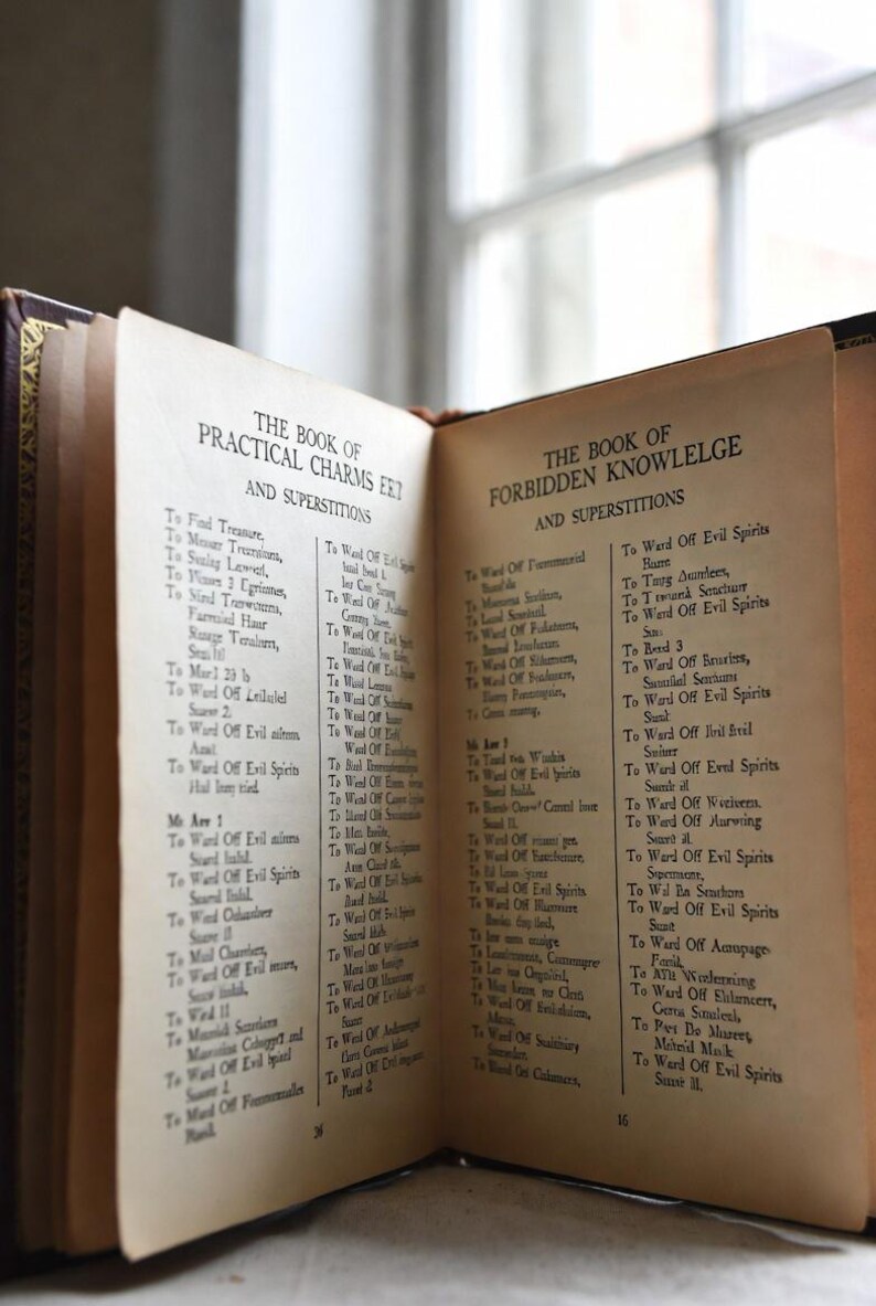 Pu&ograve; includere: Un libro aperto con pagine invecchiate, che mostra il testo di "The Book of Practical Charms" e "The Book of Forbidden Knowlege". La copertina del libro &egrave; scura con dettagli dorati. Il testo &egrave; in nero.