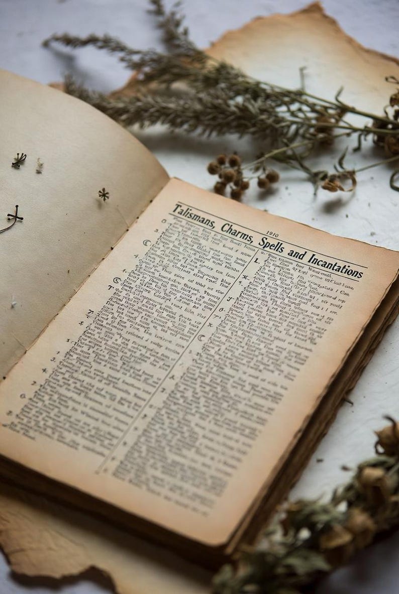 Pu&ograve; includere: Un libro antico aperto con pagine ingiallite. Il titolo "Talismans, Charms, Spells and Incantations" &egrave; visibile. Erbe secche ed elementi decorativi circondano il libro, creando un'estetica mistica.