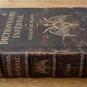 May include: An antique brown leather-bound book titled "Dictionnaire Infernal" by Collin de Plancy, with gold embossed lettering and a winged creature design. The spine shows the year 1863.