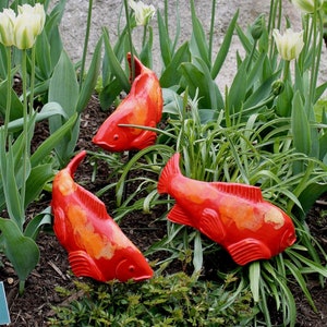 Puede incluir: Tres esculturas de peces de cerámica roja y naranja se colocan en un jardín con tulipes blancas. Los peces están dispuestos en forma de triángulo, con las colas apuntando hacia el centro.