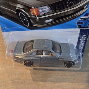 May include: A die-cast model car in a clear plastic package. The car is a silver coupe with black wheels and a white stripe on the tires. The packaging has a blue and white design with the number "2/5".