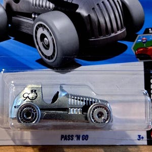 May include: A silver die-cast toy car with black wheels, packaged in a clear plastic case. The car has a vintage design with the text "PASS 'N GO" and a logo with a top hat. The packaging also indicates the car is part of a 1/5 series.