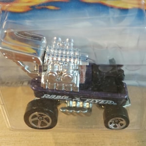 May include: A purple and silver toy truck with large chrome engine components. The toy truck has black wheels and the words "Radio Flyer" printed on the side. The toy is packaged in a clear plastic container.