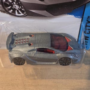 May include: A silver toy car with a futuristic design. The car has a red tinted window and black wheels. The rear of the car has a fin with multiple circular details. The car is packaged in a clear plastic container.