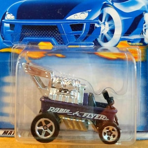 May include: A purple Radio Flyer wagon toy car in original blister packaging. The wagon features a large chrome engine block mounted on top, a black seat, and four black wheels with silver rims. The packaging displays a blue race car graphic.