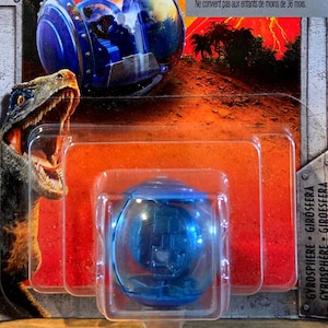 May include: A blue plastic gyrosphere toy encased in clear blister packaging. The cardboard backing features a graphic of a dinosaur with an open mouth and a jungle scene with a volcano. The text Gyrosphere is printed on the side.