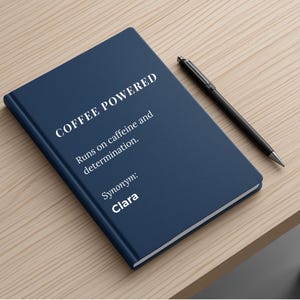 Pode incluir: Um caderno azul marinho com o texto "COFFEE POWERED" e a frase "Funciona com cafeína e determinação". Uma caneta preta repousa sobre a superfície de madeira ao lado do caderno. O sinónimo "Clara" também está impresso.