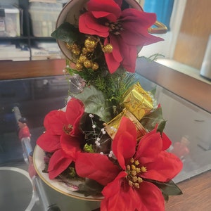 Peut inclure: Une tasse à thé blanche renversée avec une fleur de poinsettia rouge qui en sort. La tasse à thé est posée sur une soucoupe avec d'autres poinsettias et des cadeaux emballés dans du papier doré.