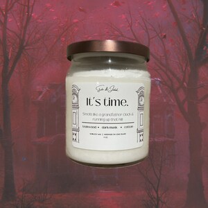 May include: A white candle jar with a copper lid. The label reads "It's time." and "Smells like a grandfather clock & running up that hill." The candle is scented with teakwood, dark musk, and cotton. It is 100% soy wax and handmade on Long Island.