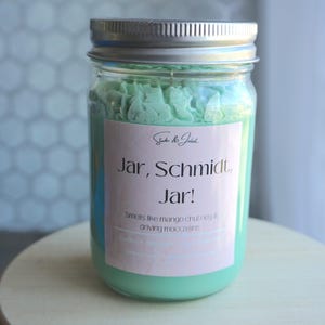 May include: A glass jar with a silver lid filled with a light green scented candle. The label reads "Shake & Jaket Jar, Schmidt, Jar! Smells like mango chutney & driving moccasins."