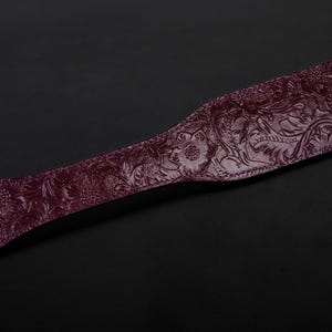 May include: A dark purple leather paddle with an embossed floral design. The paddle has a black strap and a gold metal ring.