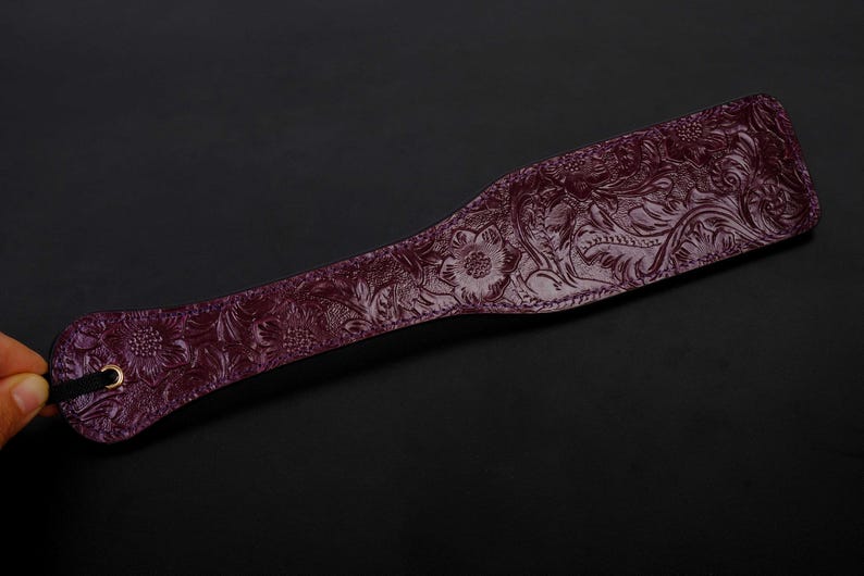 May include: A dark purple leather paddle with an embossed floral design. The paddle has a black strap and a gold metal ring.