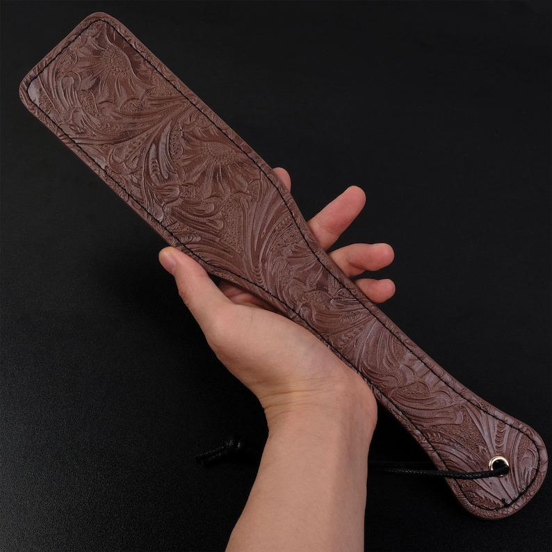 Puede incluir: Una paleta de cuero marr&oacute;n con un dise&ntilde;o floral en relieve. La paleta tiene un borde cosido y un peque&ntilde;o anillo de metal en el extremo del mango.