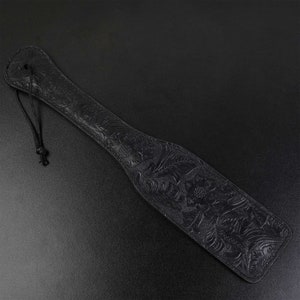 Puede incluir: Una paleta de cuero negro con un dise&ntilde;o floral en relieve. La paleta tiene un lazo en la parte superior para colgarla.
