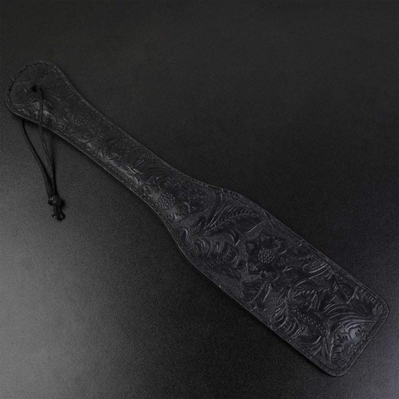 Puede incluir: Una paleta de cuero negro con un dise&ntilde;o floral en relieve. La paleta tiene un lazo en la parte superior para colgarla.