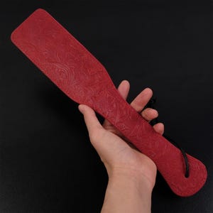Puede incluir: Una paleta de cuero roja con un dise&ntilde;o floral en relieve. La paleta tiene un cord&oacute;n negro.