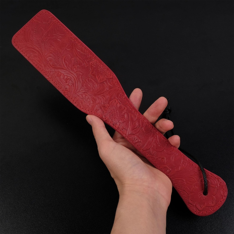 Puede incluir: Una paleta de cuero roja con un dise&ntilde;o floral en relieve. La paleta tiene un cord&oacute;n negro.