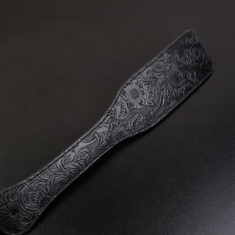 Puede incluir: Una paleta de cuero negro con un dise&ntilde;o floral en relieve. La paleta es larga y estrecha con un extremo redondeado.