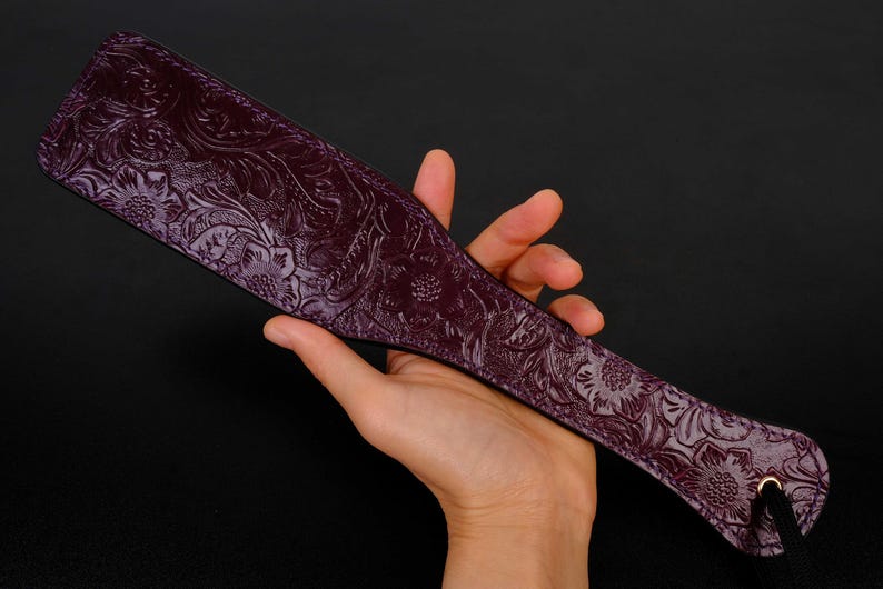 May include: A purple leather paddle with an embossed floral design. The paddle has a black cord attached to the end.