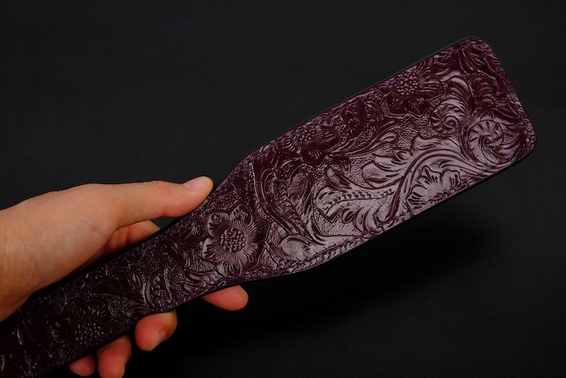 May include: A dark purple leather paddle with an intricate floral pattern embossed on the surface.