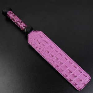 Puede incluir: Una paleta de cuero rosa con un mango trenzado negro. La paleta tiene un patrón de piel de cocodrilo.