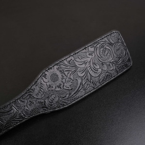Puede incluir: Una paleta de cuero negro con un dise&ntilde;o floral en relieve. La paleta tiene una superficie lisa y pulida y un borde ligeramente redondeado.