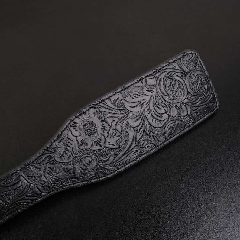 Puede incluir: Una paleta de cuero negro con un dise&ntilde;o floral en relieve. La paleta tiene una superficie lisa y pulida y un borde ligeramente redondeado.