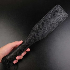 Puede incluir: Una paleta de cuero negro con un dise&ntilde;o floral en relieve. La paleta se sostiene en una mano.