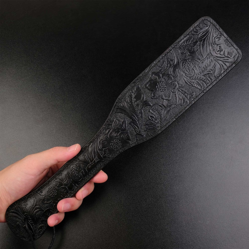Puede incluir: Una paleta de cuero negro con un dise&ntilde;o floral en relieve. La paleta se sostiene en una mano.