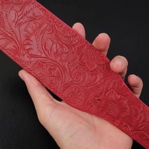May include: A red leather guitar strap with a floral embossed design. The strap is shown being held in a hand.