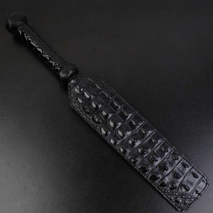 May include: A black leather paddle with a braided handle and a crocodile-embossed surface.