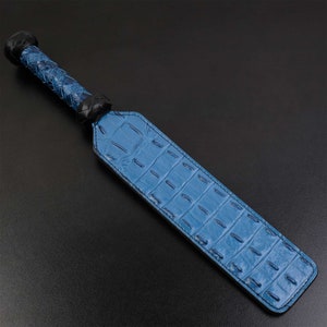 May include: A blue leather paddle with a black leather handle. The paddle has a crocodile skin pattern.
