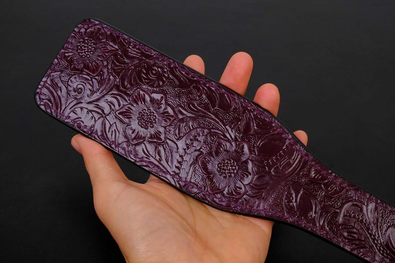 May include: A purple leather paddle with an embossed floral design. The paddle is held in a hand.
