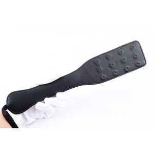 May include: A black leather paddle with a textured surface and a handle. The paddle has multiple small holes throughout the surface.