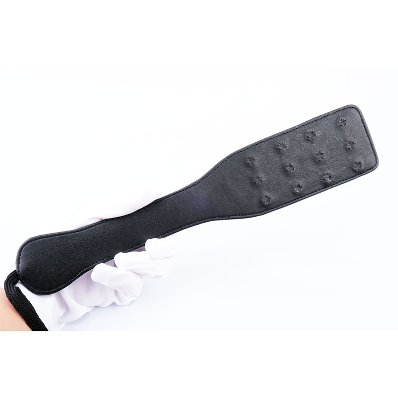 May include: A black leather paddle with a textured surface and a handle. The paddle has multiple small holes throughout the surface.