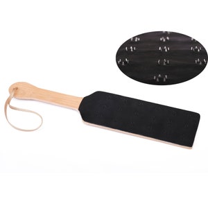 May include: A black leather paddle with a wooden handle and a strap. The paddle has a pattern of small metal studs on the surface.