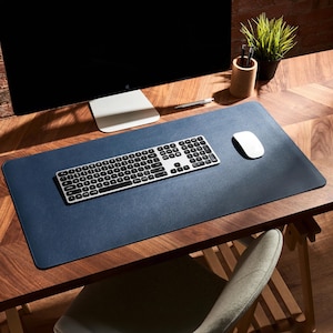 May include: A rectangular navy blue desk mat with stitched edges, positioned on a wooden surface. The mat features a smooth, flat surface supporting a silver wireless keyboard with black keys and a white computer mouse placed to the right side.