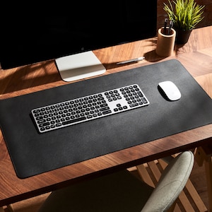 May include: A black leather desk mat with a silver keyboard and a white wireless mouse. The mat is on a wooden desk with a computer monitor and a pencil holder.