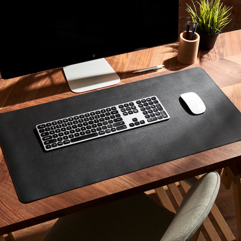 May include: A black leather desk mat with a silver keyboard and a white wireless mouse. The mat is on a wooden desk with a computer monitor and a pencil holder.