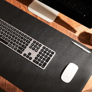 May include: A black leather desk mat with a silver keyboard, a white wireless mouse, and a silver pen on a wooden surface.