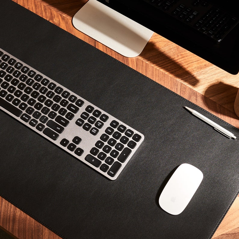 May include: A black leather desk mat with a silver keyboard, a white wireless mouse, and a silver pen on a wooden surface.