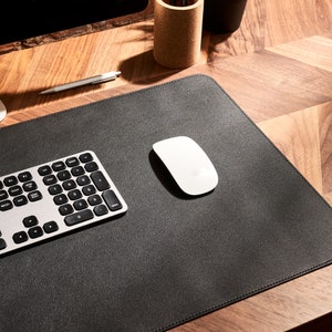 May include: A black leather desk mat with a silver keyboard and a white wireless mouse. The mat is on a wooden desk.