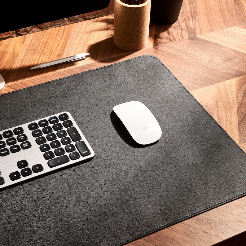 May include: A black leather desk mat with a silver keyboard and a white wireless mouse. The mat is on a wooden desk.