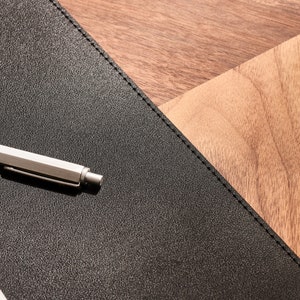 May include: A silver pen lies on a black leather desk pad with a wood grain background.