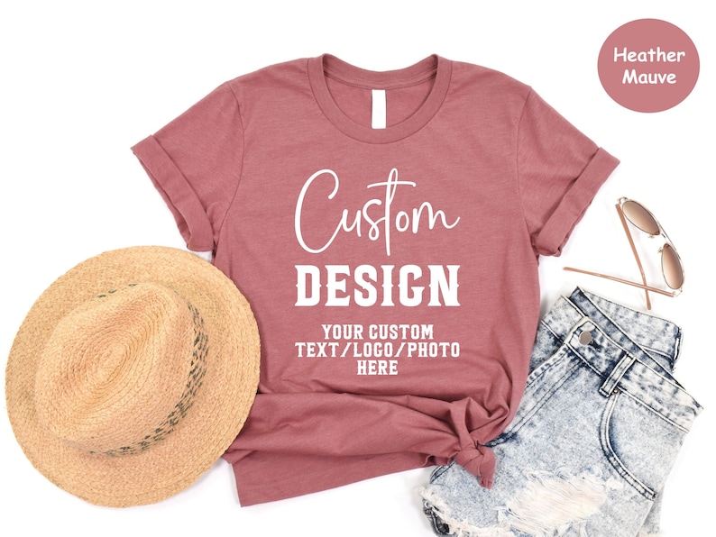 May include: A heather mauve t-shirt with the text "Custom Design" and "Your Custom Text/Logo/Photo Here" in white. The t-shirt is laid flat on a white surface with a straw hat and denim shorts.