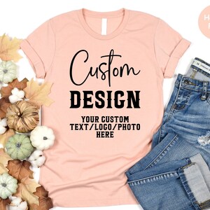 May include: A heather peach colored t-shirt with the text "Custom Design" and "Your Custom Text/Logo/Photo Here" printed on it. The t-shirt is laying on a white surface with fall leaves and gourds around it.