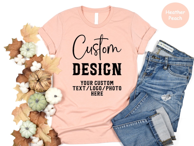 May include: A heather peach colored t-shirt with the text "Custom Design" and "Your Custom Text/Logo/Photo Here" printed on it. The t-shirt is laying on a white surface with fall leaves and gourds around it.