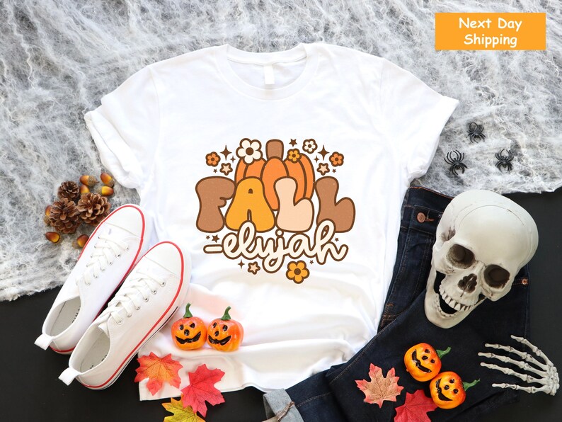 May include: White t-shirt featuring a retro design with the words "FALL eliyah" in brown, orange, and beige, with pumpkin and flower illustrations. The shirt is styled with white trainers, jeans, and Halloween decorations.